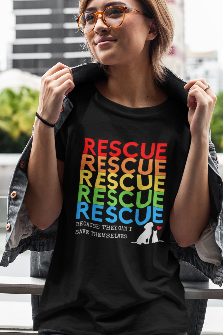 A woman wearing a black “Rescue – Because They Can’t Save Themselves” rainbow animal rescue shirt, standing outdoors in the city. This colorful cat and dog rescue T-shirt promotes TNR, fostering, and animal rescue awareness — perfect for volunteers and compassionate pet lovers.