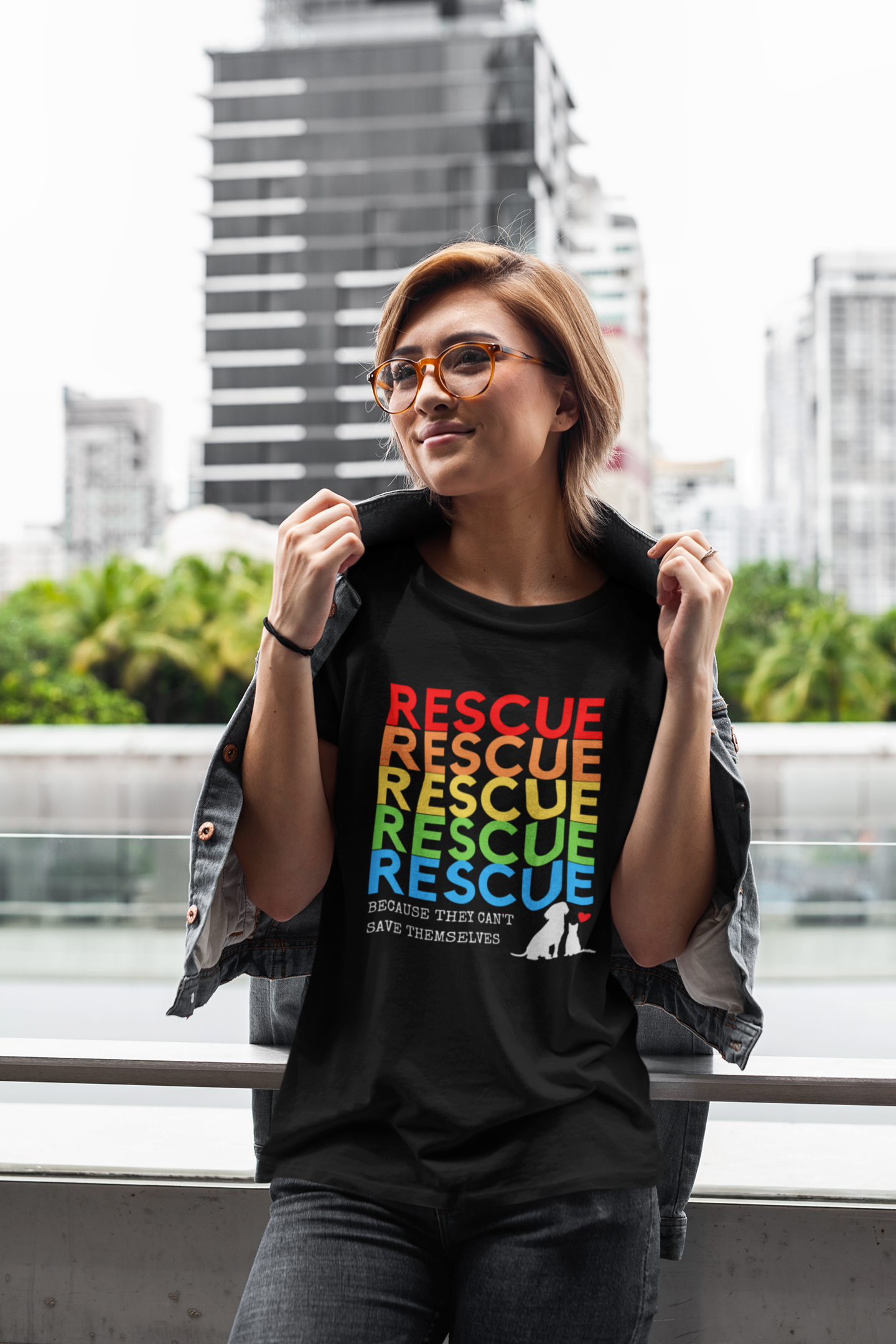 A woman wearing a black “Rescue – Because They Can’t Save Themselves” rainbow animal rescue shirt, standing outdoors in the city. This colorful cat and dog rescue T-shirt promotes TNR, fostering, and animal rescue awareness — perfect for volunteers and compassionate pet lovers.