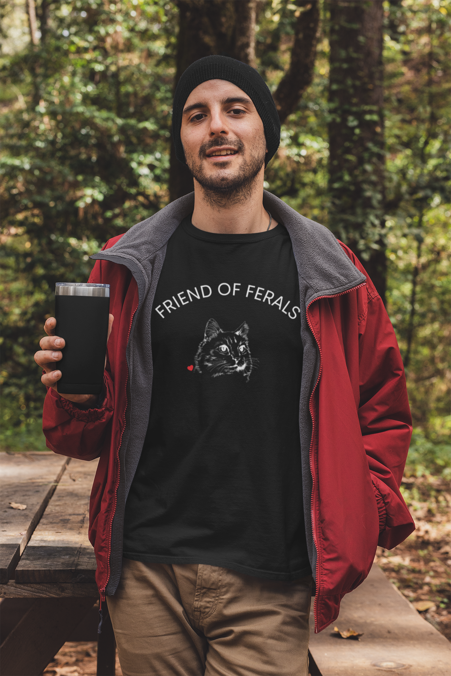 A man wearing a black “Friend of Ferals” TNR awareness shirt featuring a detailed cat illustration and red heart, standing outdoors in the woods. This feral cat rescue shirt supports animal rescue, TNR, and community cat advocates — perfect gift for cat lovers and volunteers.