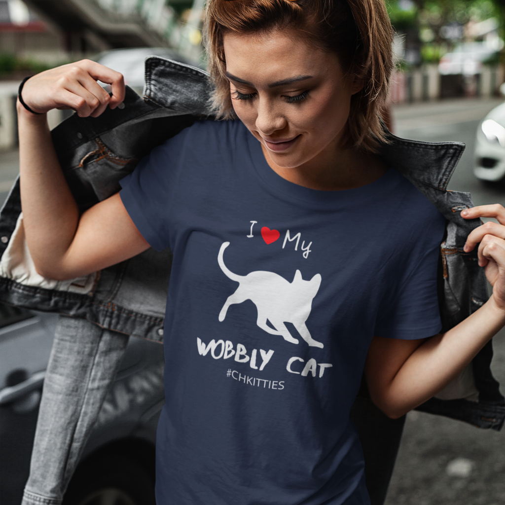 Lifestyle mockup of woman wearing a Navy t-shirt with a white silhouette of a wobbly cat and text reading ‘I Love My Wobbly Cat #CHKITTIES.’ Special-needs CH cerebellar hypoplasia cat awareness shirt 