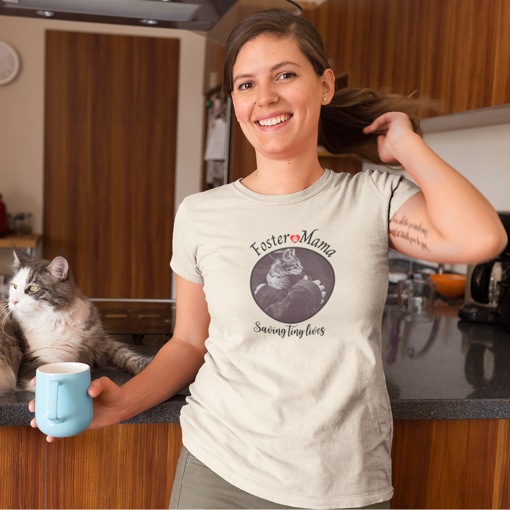 Person in kitchen wearing a t-shirt with 'Foster Mama Saving Tiny Lives' design featuring a kitten and person holding tabby foster kitten.